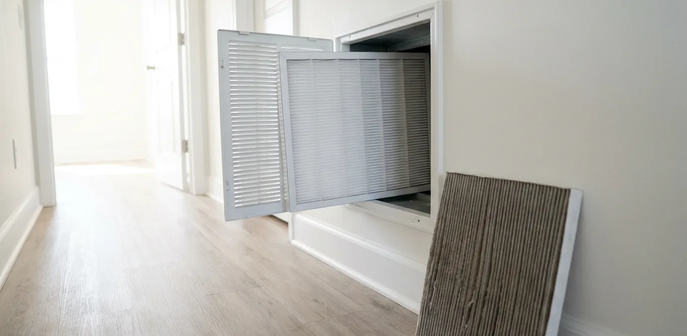 An open return-air grille in a hallway with a clean white pleated filter going in and the dusty old filter on the floor